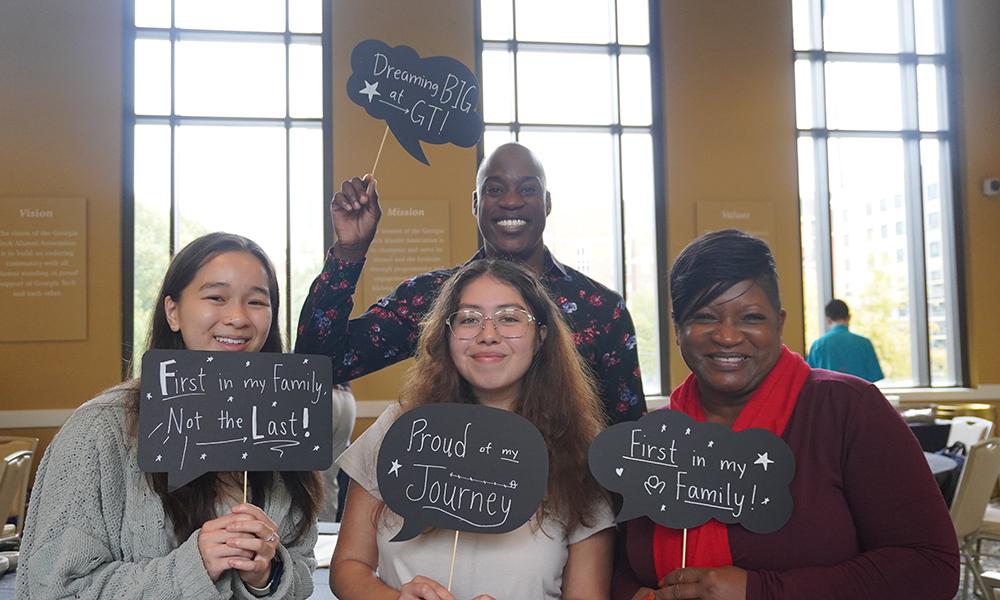 Associate Director of First-Generation Student Initiatives Charmaine Troy (far right) holds up sign that reads "First in my family" with students and MC from First-Generation Symposium.